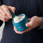 A person dips a shaving brush into a container of Barberino's Bergamot and Neroli Shave Soap, holding the brush in one hand and the luxury grooming soap in the other.