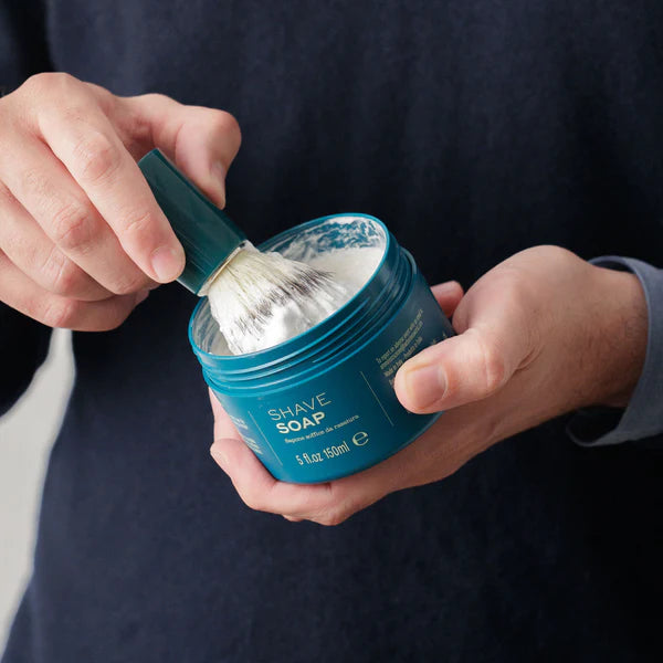 A person dips a shaving brush into a container of Barberino's Bergamot and Neroli Shave Soap, holding the brush in one hand and the luxury grooming soap in the other.