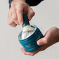 A person uses the Barberino's Cruelty Free Shaving Brush to swirl lathering shaving soap in a container, creating a rich lather for a traditional shave.