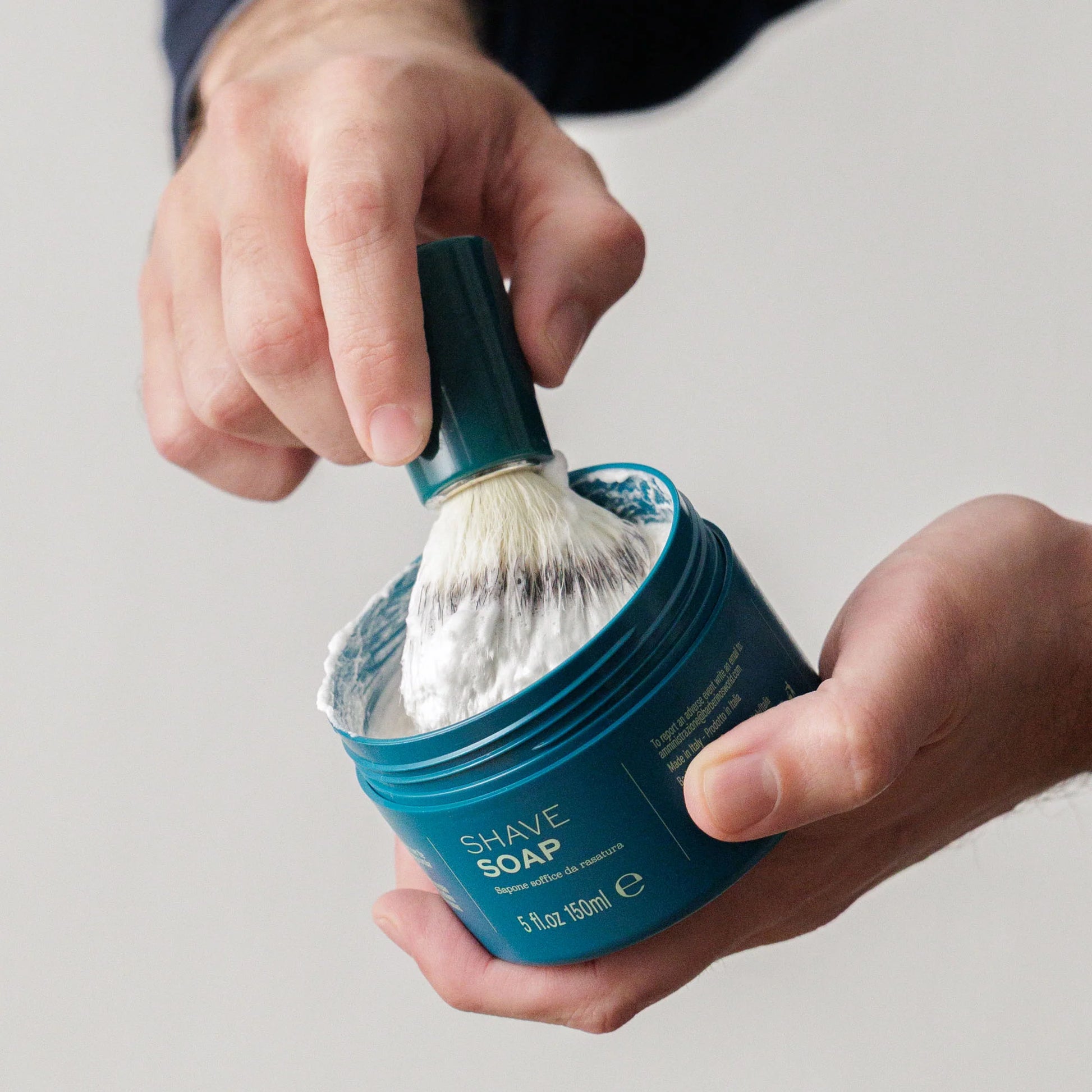 A person uses the Barberino's Cruelty Free Shaving Brush to swirl lathering shaving soap in a container, creating a rich lather for a traditional shave.