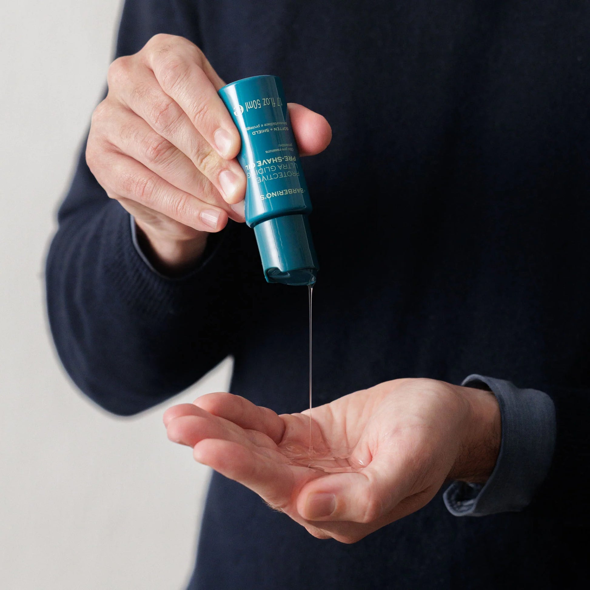 A person dispenses Barberino's Protective Ultra Gliding Pre-Shave Oil from a blue bottle into their palm while wearing a dark long-sleeve shirt, preparing for a smoother shave.