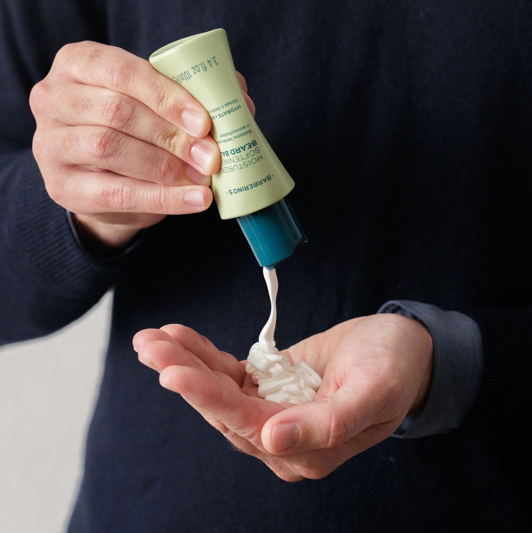 A person dispenses Barberino's Moisturizing Beard Balm from a green tube into their palm while wearing a dark long-sleeve shirt—ideal for moisturizing beard care.