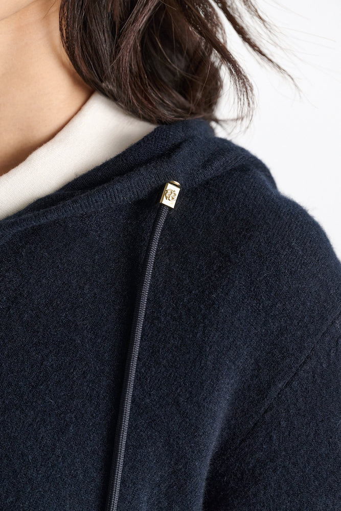 Close-up of a person wearing the Dorothee Schumacher Modern Statements Pullover, a cropped dark navy hoodie with a gold metal-tipped drawstring. Only part of their face, shoulder, and hair is shown.