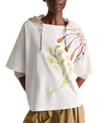 A person wears the Dorothee Schumacher Daydream Flower Shirt—a relaxed cotton tee with dropped shoulders and a floral print—paired with a beige drawstring hood and light brown pants.