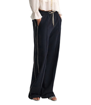 A person wears Dorothee Schumacher’s Casual Attraction Pants—relaxed fit, dark navy with white piping and an elastic drawstring waist—paired with a white long-sleeved blouse and sandals.