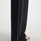 A person stands on a light surface wearing Dorothee Schumacher's Casual Attraction Pants—black wool joggers with an elastic waist and white piping—paired with metallic silver open-toe sandals.