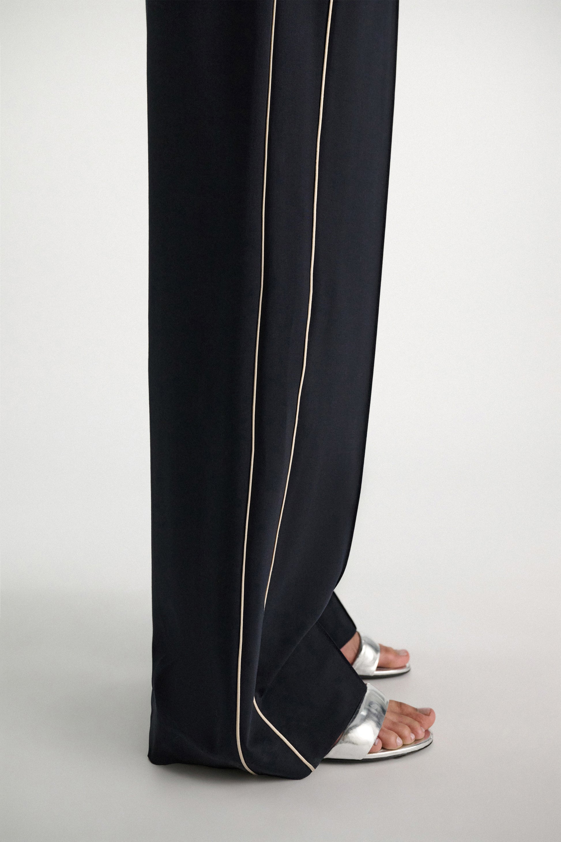 A person stands on a light surface wearing Dorothee Schumacher's Casual Attraction Pants—black wool joggers with an elastic waist and white piping—paired with metallic silver open-toe sandals.
