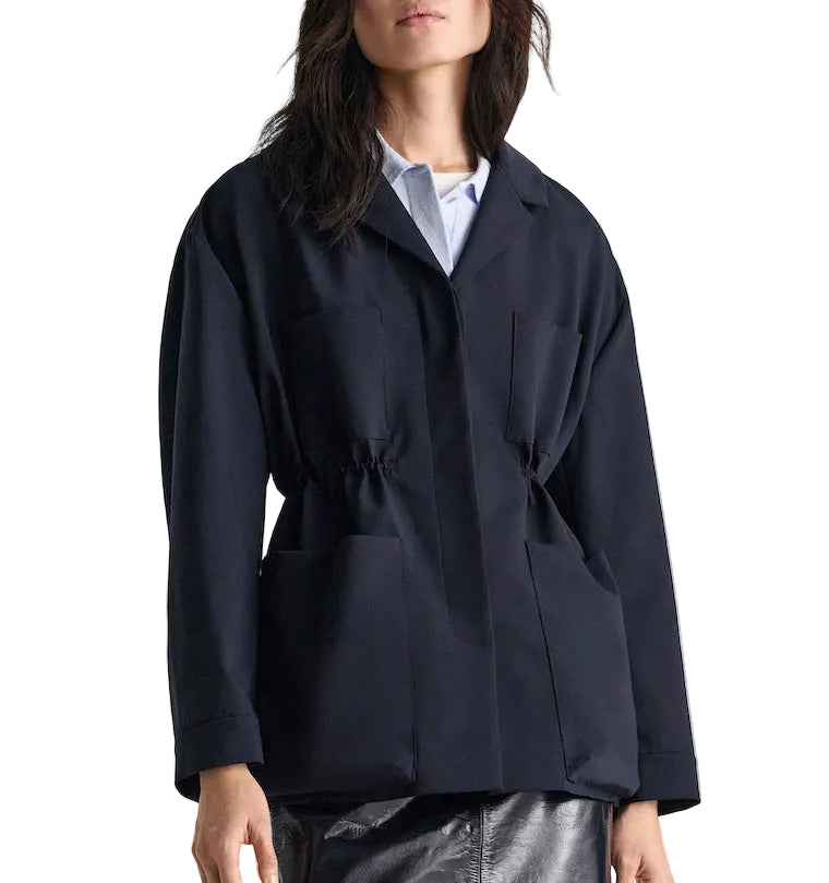 A woman styles the Dorothee Schumacher Casual Attraction Jacket—a loose-fitting black jacket with large pockets and a drawstring waist—over a light blue shirt and metallic silver skirt for an effortlessly chic modern look.