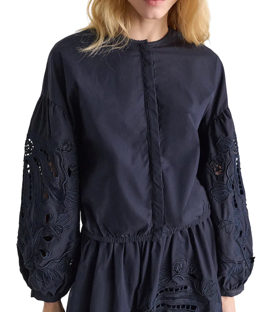 A woman wears the Dorothee Schumacher Embroidered Beauty Blouse, featuring black fabric, long sleeves, floral embroidery, and cutout details on the sleeves and waist.
