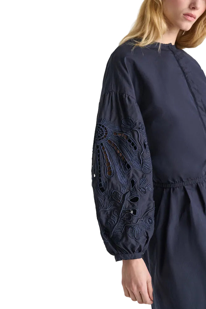 A woman models the Dorothee Schumacher Embroidered Beauty Blouse, a navy blue cotton top with floral embroidery on long sleeves, standing against a white background.