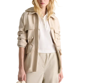 A woman wears the Dorothee Schumacher Emotional Essence Jacket in beige, unbuttoned over a white collared shirt and beige pants, her left sleeve pushed up and hair partly covering her face.