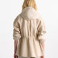 A person with long blonde hair is seen from the back wearing the Dorothee Schumacher Emotional Essence Jacket—light beige with a hood, gathered sleeves, adjustable drawstring waist, and matching pants—against a plain background.