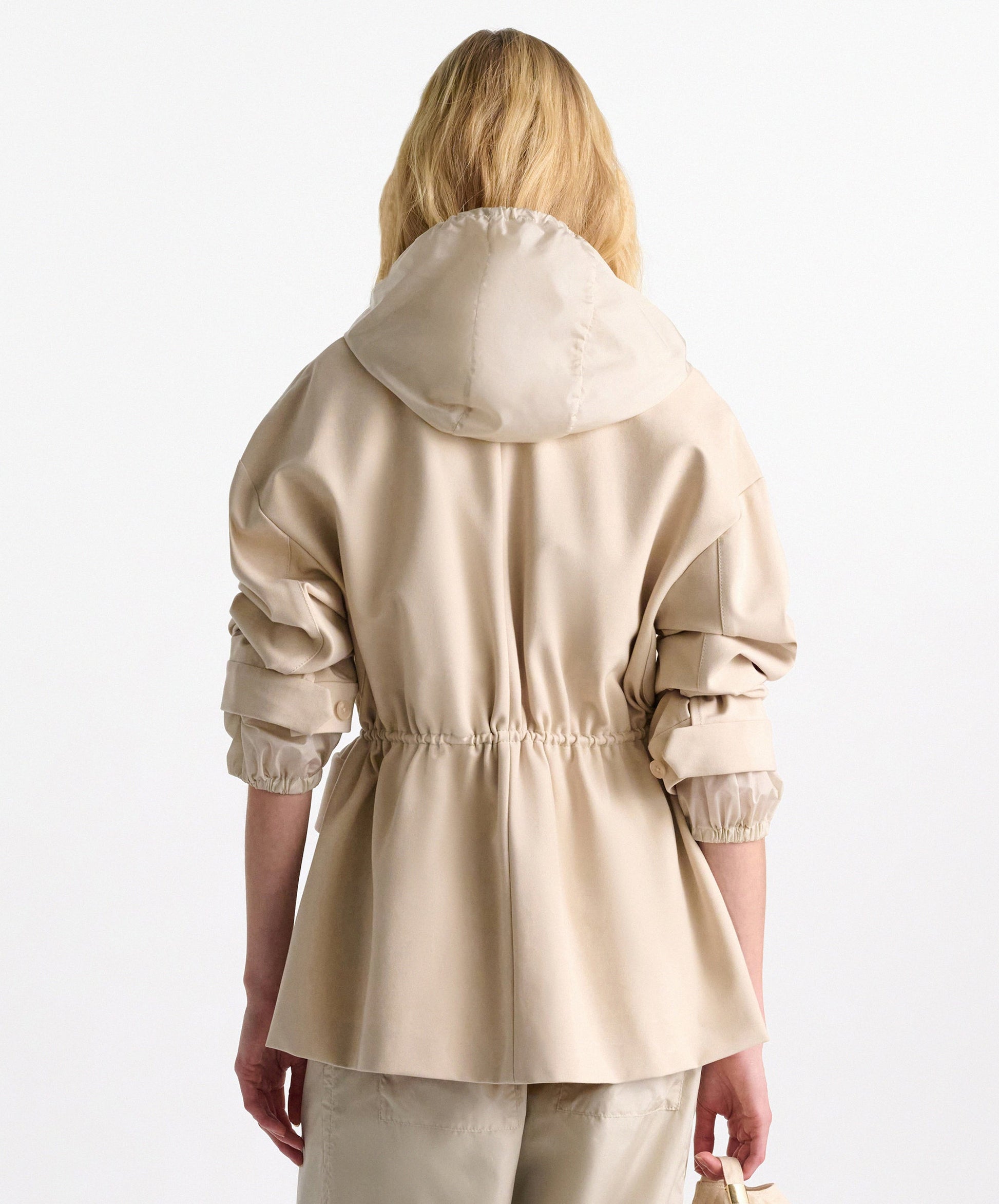 A person with long blonde hair is seen from the back wearing the Dorothee Schumacher Emotional Essence Jacket—light beige with a hood, gathered sleeves, adjustable drawstring waist, and matching pants—against a plain background.