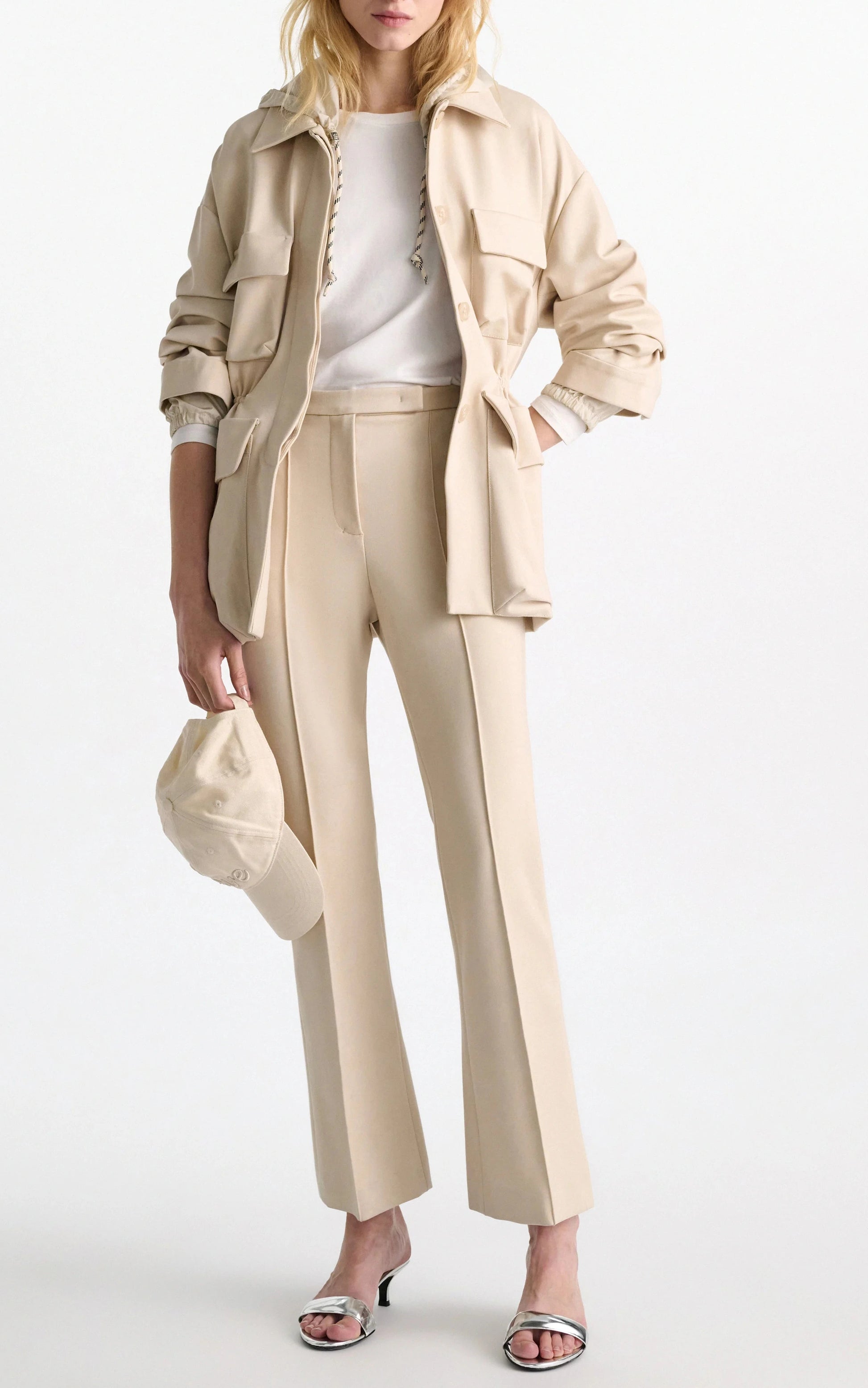 A woman wears Dorothee Schumacher's Emotional Essence Pants with a beige utility jacket, white top, heeled sandals, and holds a beige baseball cap in her left hand.