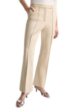 A person wears Dorothee Schumacher’s Emotional Essence Pants—beige, cropped, tailored with front seams—paired with a white top and silver heeled sandals, standing with one hand in a pocket.