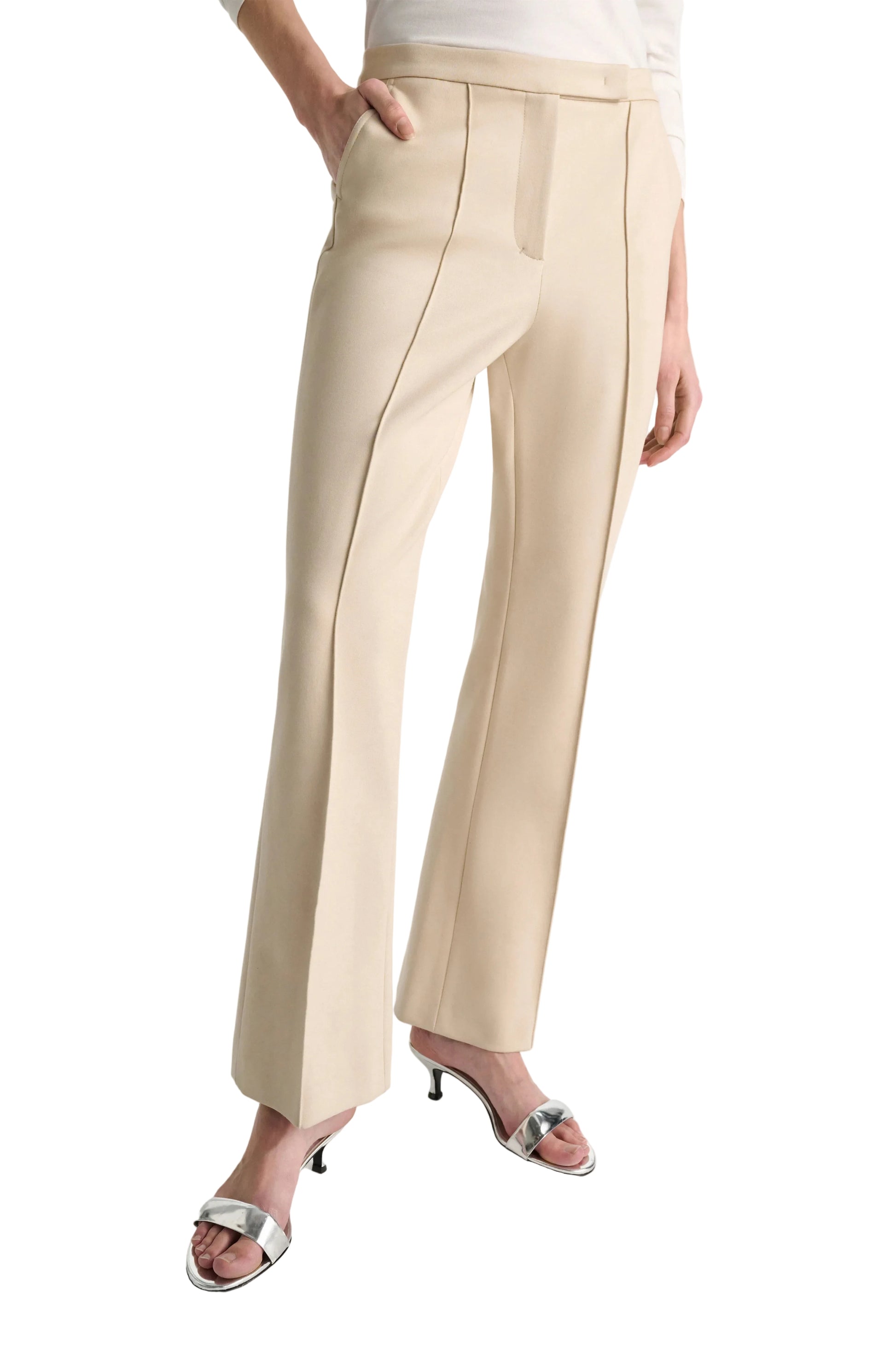 A person wears Dorothee Schumacher’s Emotional Essence Pants—beige, cropped, tailored with front seams—paired with a white top and silver heeled sandals, standing with one hand in a pocket.