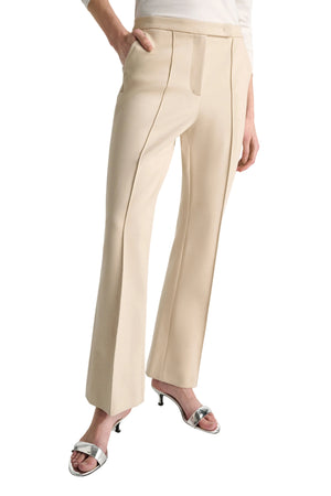 A person wears Dorothee Schumacher’s Emotional Essence Pants—beige, cropped, tailored with front seams—paired with a white top and silver heeled sandals, standing with one hand in a pocket.