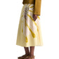 A person wears the Dorothee Schumacher Floral Power Skirt—a yellow signature print cotton poplin midi-skirt with a fitted waist—paired with a brown jacket and flat sandals, standing against a plain background.