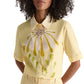 Woman wears the Floral Power Blouse by Dorothee Schumacher, a light yellow cotton poplin top with a bold front floral design and a removable lace collar for versatile styling.