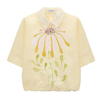 The Floral Power Blouse by Dorothee Schumacher is a light yellow cotton poplin top featuring a removable lace collar and a bold pastel pink, yellow, and green floral design on the front.