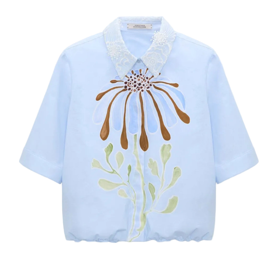 Dorothee Schumacher’s Floral Power Blouse in light blue cotton poplin features short sleeves, a removable lace collar, and bold abstract floral prints in brown, blue, and green on the front.
