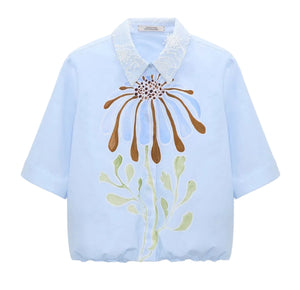 Dorothee Schumacher’s Floral Power Blouse in light blue cotton poplin features short sleeves, a removable lace collar, and bold abstract floral prints in brown, blue, and green on the front.