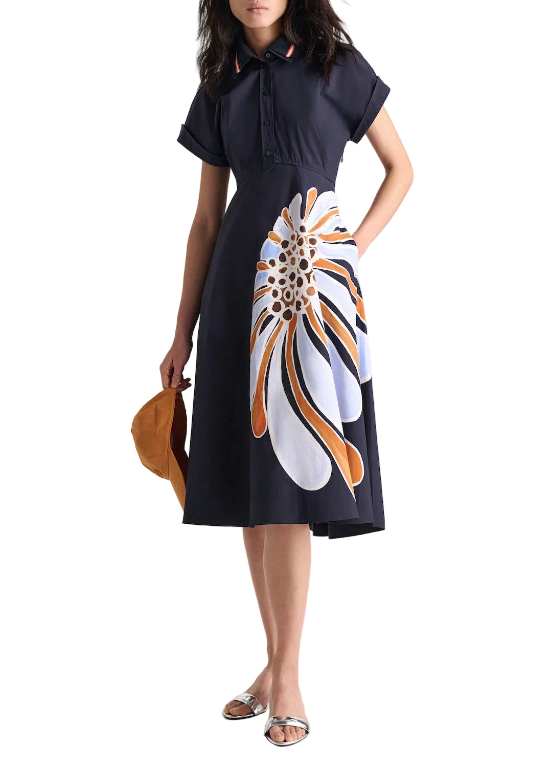 A woman in the Dorothee Schumacher Floral Power Dress, featuring a navy blue cotton poplin fabric, polo collar, and abstract floral print skirt, holds a brown hat and wears silver flat sandals.