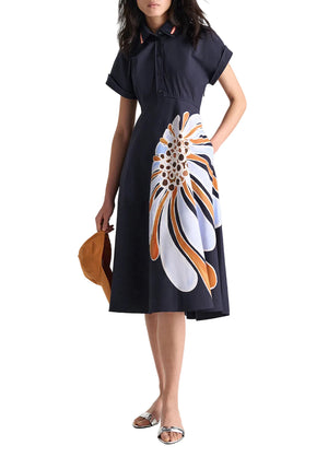 A woman in the Dorothee Schumacher Floral Power Dress, featuring a navy blue cotton poplin fabric, polo collar, and abstract floral print skirt, holds a brown hat and wears silver flat sandals.