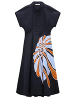 The Floral Power Dress by Dorothee Schumacher is a navy cotton poplin piece with a polo collar, short sleeves, buttons, and a bold floral print in white, blue, and orange on one side of the skirt.