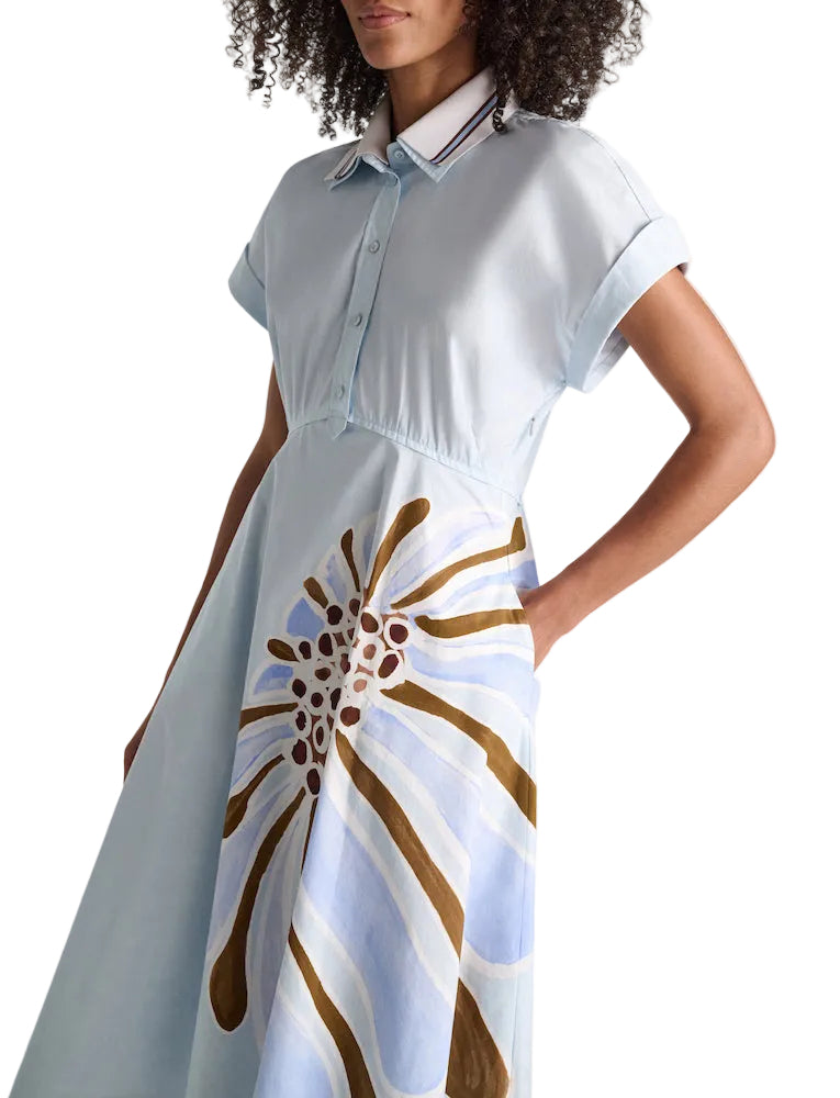A woman wears the Dorothee Schumacher Floral Power Dress, a light blue cotton poplin piece with a polo collar and bold abstract brown and blue floral print on the skirt.