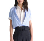 A woman wears the Powerful Ease Cropped Blouse by Dorothee Schumacher layered under a light blue open shirt, styled with high-waisted black pants and a wide black belt.
