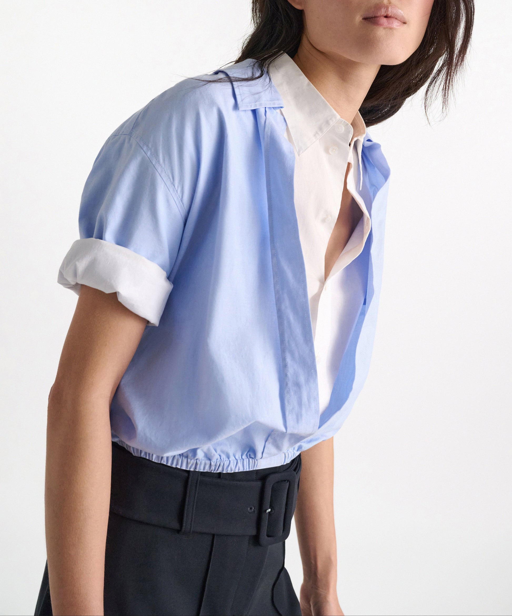 A woman wears the Dorothee Schumacher Powerful Ease Cropped Blouse in light blue over a white oxford shirt, paired with high-waisted belted black pants.