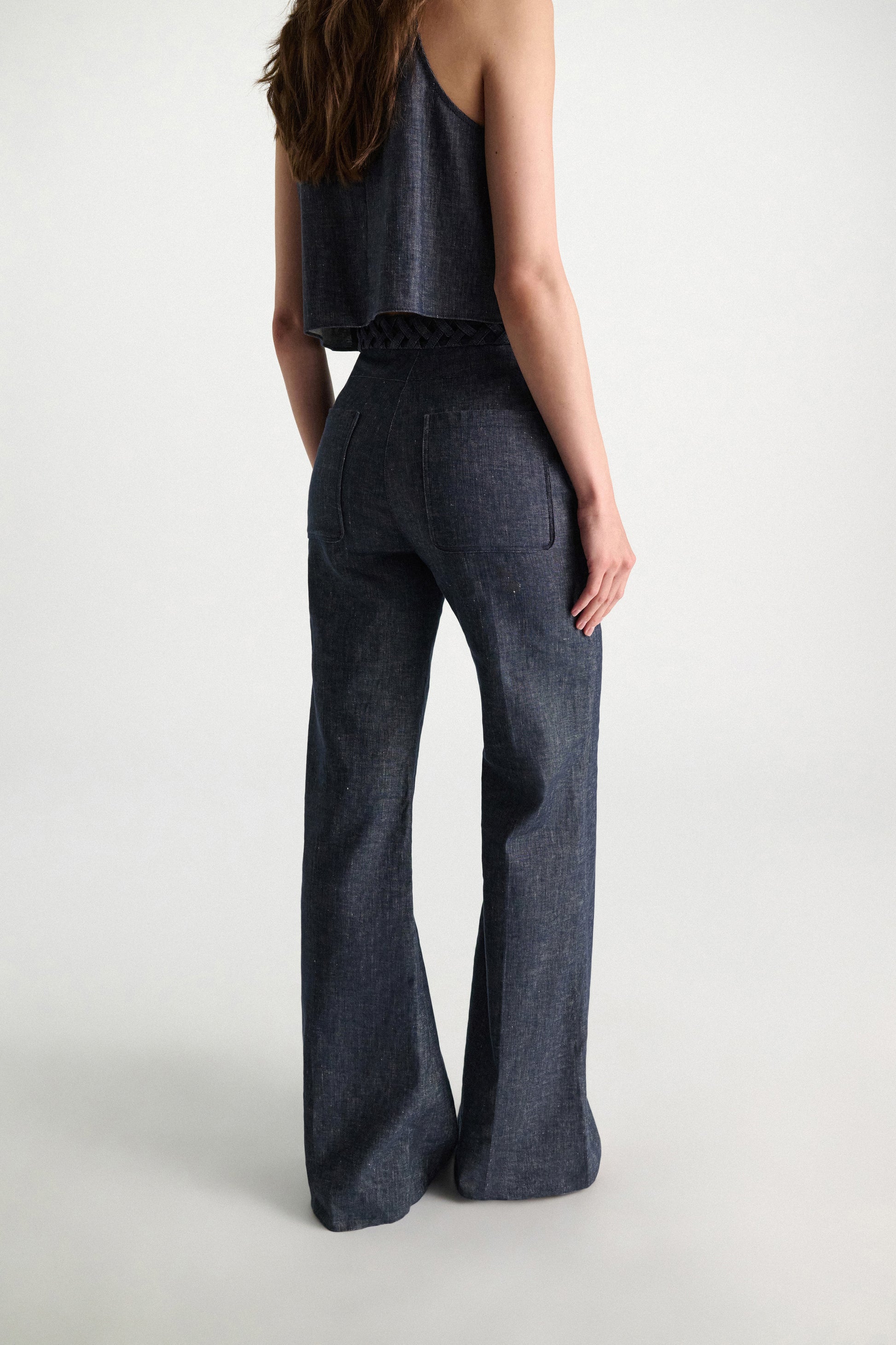 A woman is seen from behind wearing Dorothee Schumacher's Fluid Denim Pants—high-waisted, wide-leg, dark denim with subtle braid detailing—paired with a sleeveless matching top against a plain light background.