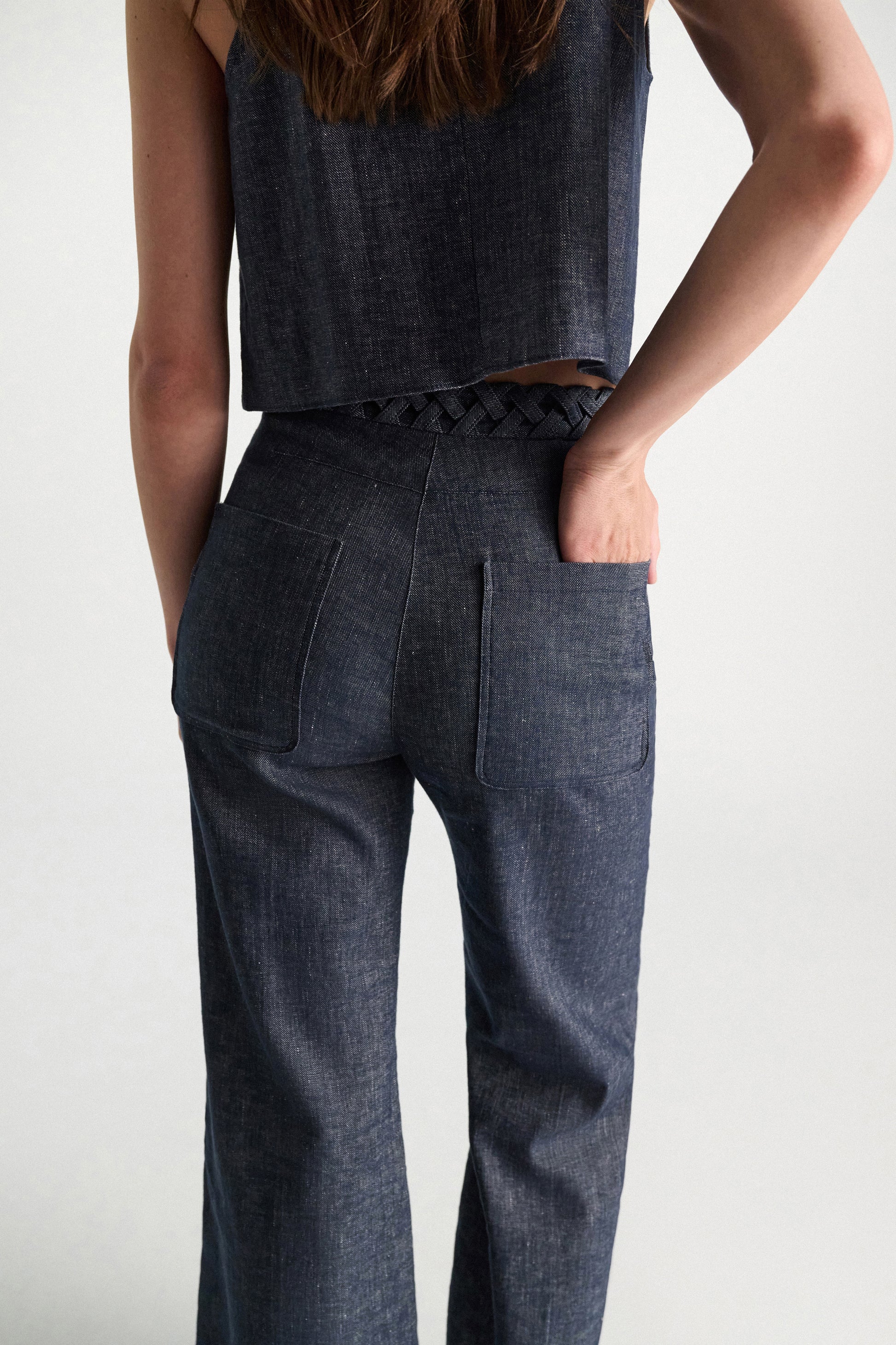 A person in Dorothee Schumacher's Fluid Denim Pants and a sleeveless dark denim braided-detail top, seen from the back with one hand in their pocket.