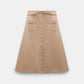 The Dorothee Schumacher Luxury Layer Skirt is a beige flared design in technical cotton, featuring a front button closure, belted patch pockets with zippers, and a matching waist belt, shown against a white background.