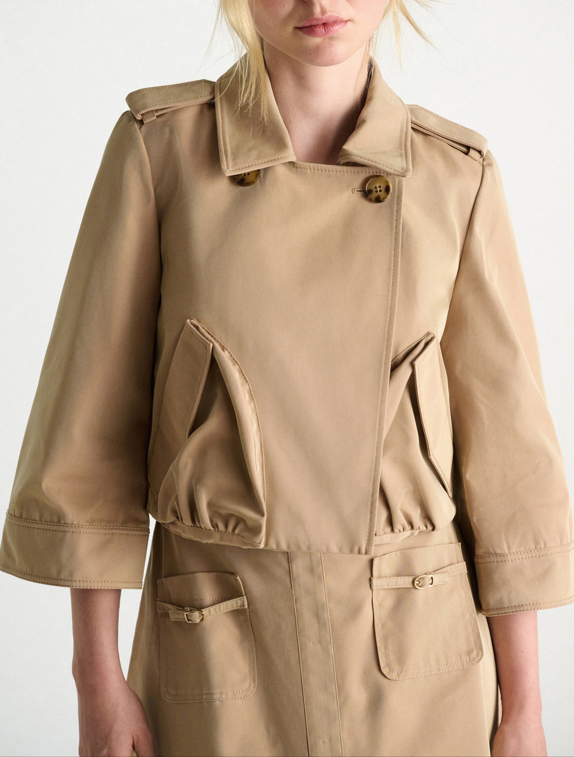 A person wears the Dorothee Schumacher Luxury Layer Jacket in beige, featuring 3/4 sleeves, large angled front pockets, and button details. The photo is cropped from chin to mid-thigh.