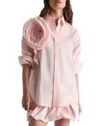 A woman wears the Dorothee Schumacher Striped Power Blouse—an oversized light pink shirt with rolled sleeves and a fabric flower on the left shoulder—paired with a matching pink skirt.