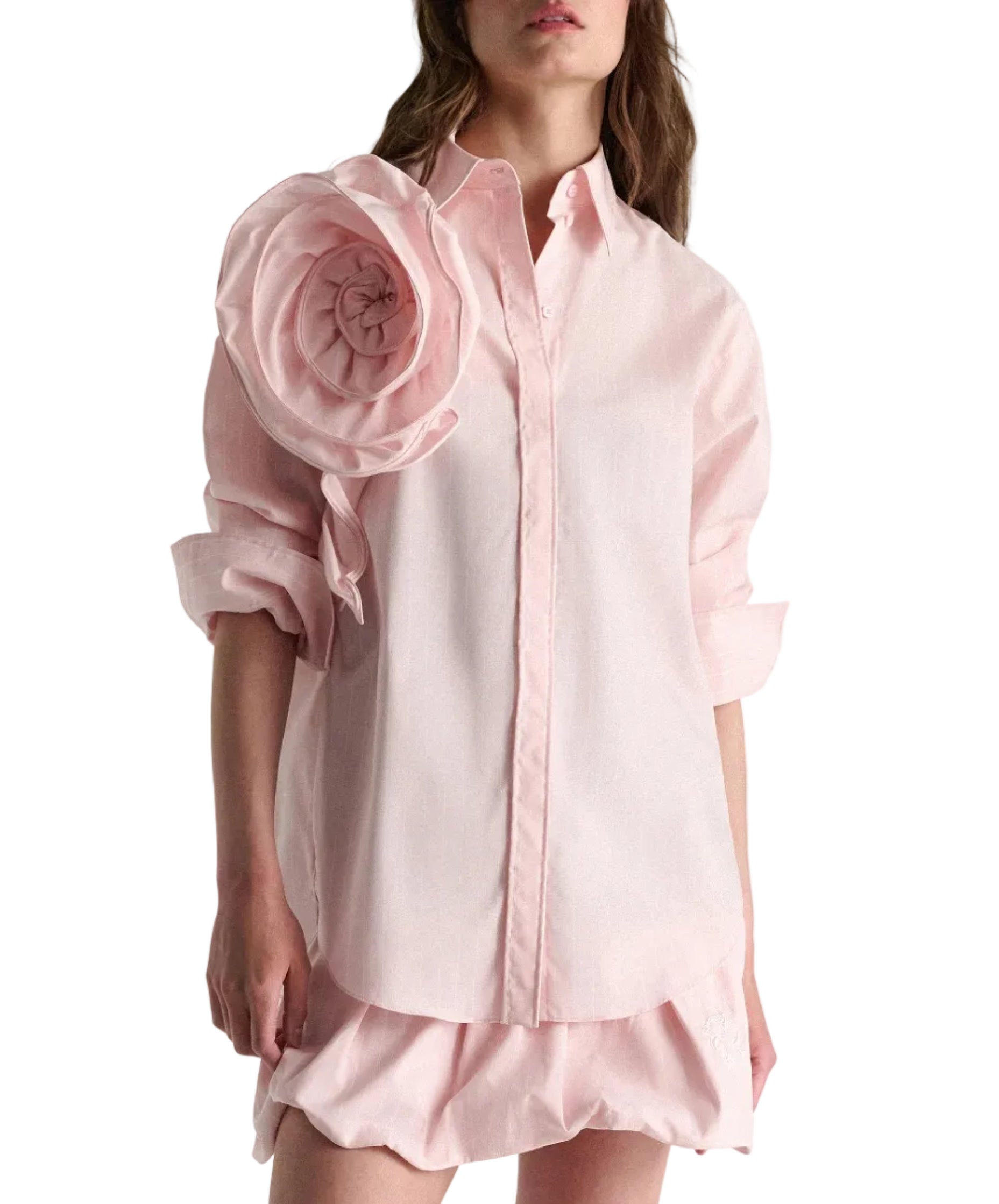 A woman wears the Dorothee Schumacher Striped Power Blouse—an oversized light pink shirt with rolled sleeves and a fabric flower on the left shoulder—paired with a matching pink skirt.