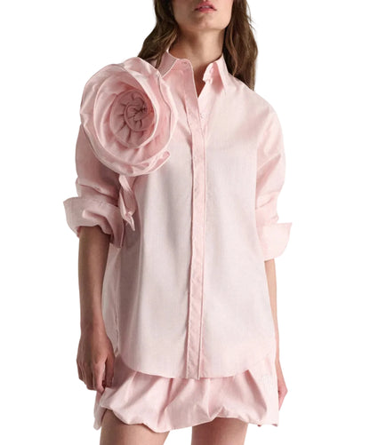A woman wears the Dorothee Schumacher Striped Power Blouse—an oversized light pink shirt with rolled sleeves and a fabric flower on the left shoulder—paired with a matching pink skirt.