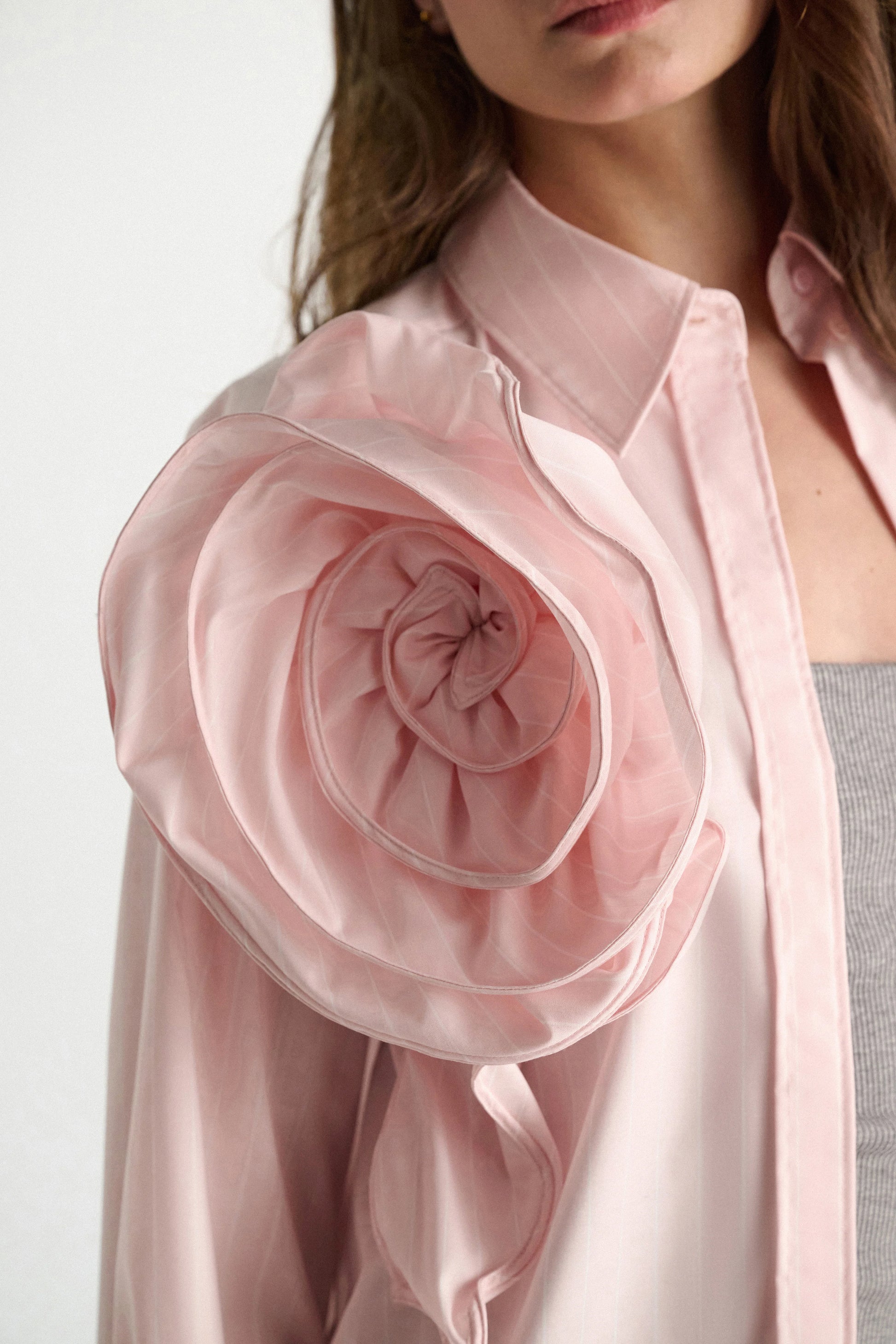 A woman wears the Dorothee Schumacher Striped Power Blouse, featuring a light pink oversized fit and a sculpted rose-shaped shoulder detail, styled over a gray ribbed top.