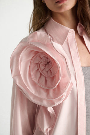 A woman wears the Dorothee Schumacher Striped Power Blouse, featuring a light pink oversized fit and a sculpted rose-shaped shoulder detail, styled over a gray ribbed top.