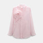 The Striped Power Blouse by Dorothee Schumacher features light pink vertical stripes, long sleeves, and a bold fabric flower brooch on the left shoulder.