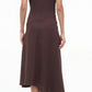 A person faces away, wearing the Staud Davina Dress—a brown, short-sleeve asymmetrical dress with a handkerchief hem—and black heeled sandals against a plain background.
