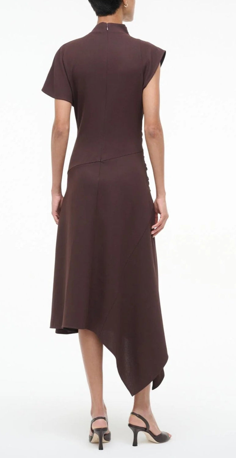 A person faces away, wearing the Staud Davina Dress—a brown, short-sleeve asymmetrical dress with a handkerchief hem—and black heeled sandals against a plain background.