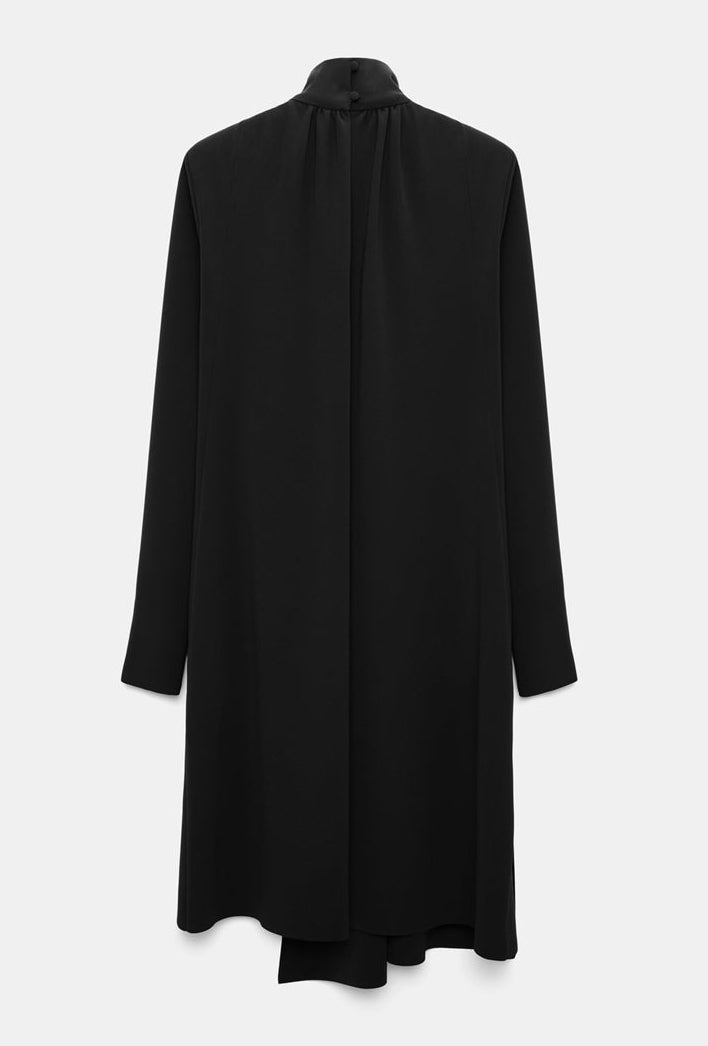 The Statement Drape Dress by Dorothee Schumacher is a long-sleeve, knee-length black dress made from luxurious Japanese crepe, showcasing a high neckline and a straight, loose fit, presented from the back.