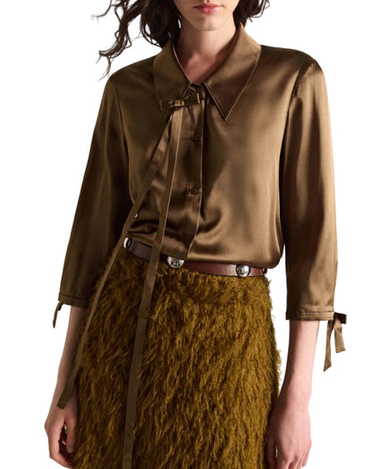 A woman wearing the Sense of Shine Blouse by Dorothee Schumacher—a brown stretch satin top featuring a removable bow tie at the neck and three-quarter sleeves—paired with a textured brown skirt and a belt adorned with round silver accents.