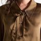 A woman wearing the Sense of Shine Blouse by Dorothee Schumacher—a brown stretch satin top featuring a pointed collar and a decorative, removable bow tie beside the buttons.