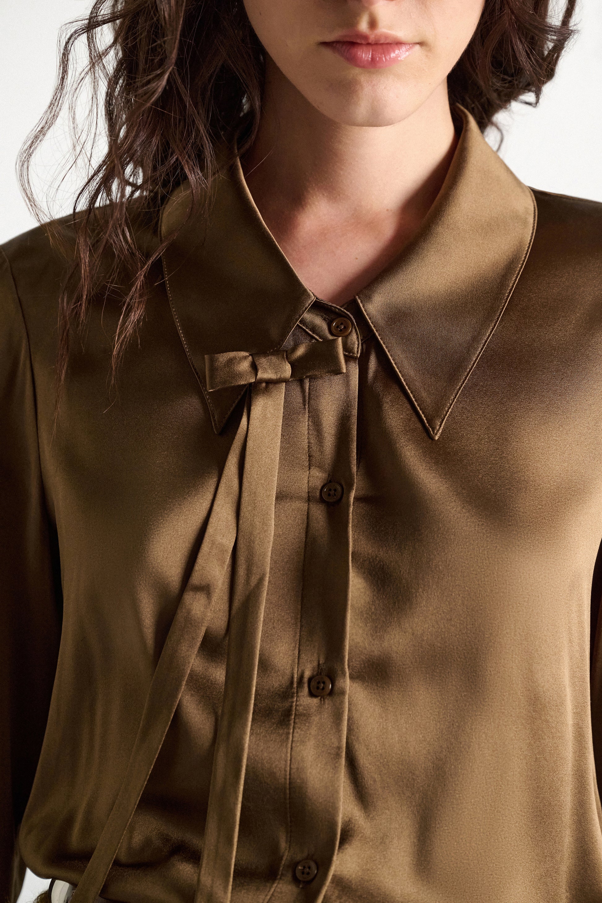 A woman wearing the Sense of Shine Blouse by Dorothee Schumacher—a brown stretch satin top featuring a pointed collar and a decorative, removable bow tie beside the buttons.