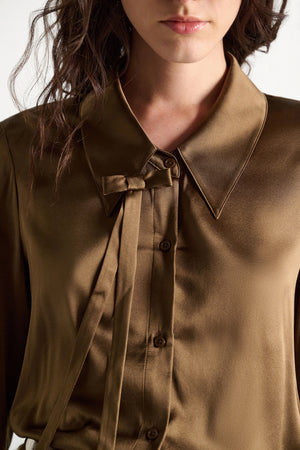 A woman wearing the Sense of Shine Blouse by Dorothee Schumacher—a brown stretch satin top featuring a pointed collar and a decorative, removable bow tie beside the buttons.