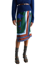 A person wearing a blue sweater, a brown belt, and the Dorothee Schumacher Cosmic Funghi Skirt—a colorful silk twill wrap skirt featuring a mushroom print and layered hemline—paired with gray socks and gray heeled sandals.